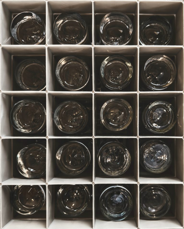 Overhead view of a box filled with identical glasses. Each glass has been neatly arranged, their uniformity creating a pattern of clear surfaces against the contrasting backdrop of the box.