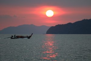 A photo of a solitary boat navigating calm waters at sunset, set against the dramatic backdrop of a mountain under a vibrant red sun
