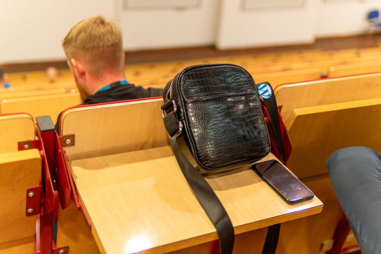 In the photo taken in the lecture hall, the foreground prominently displays a small black women’s purse, adorned with a pinned-on blue WordPress logo. In the background, a man sits with his back to the camera, dressed in a black shirt.