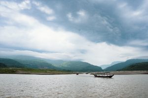Shada Pathor, Bholaganj, Sylhet, Bangladesh. Small wooden fishing boats coming out of the mouth of a river on a cloudy day. Forest covered mountains in the distance.
