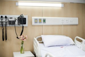 View larger photo: Hospital bed, made and unused. Flowers on a stand next to tge bed, medical equipment on the wall put away.