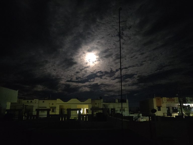 Night sky with clouds and moonlight peeking from the clouds showing houses below it.