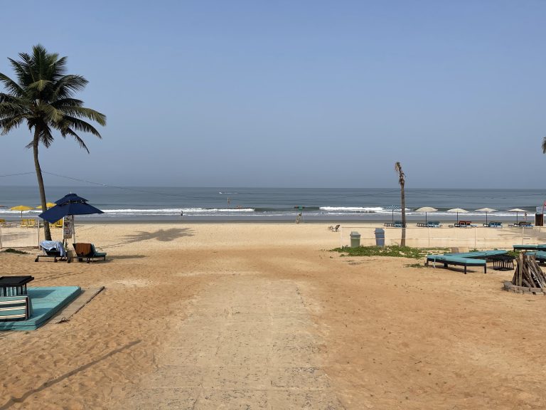 The camera view is on the beach about 100 yards from the ocean. In view are a palm tree, several beach beds, and some umbrellas. It’s a pleasant, clear day with low surf.