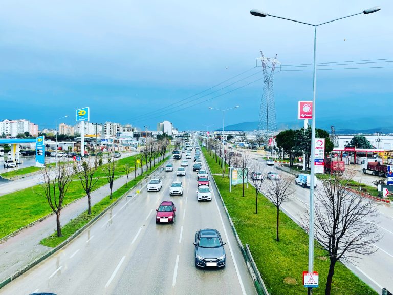 Scenic highway with light traffic, green grass dividers, and mountainous backdrop. Photo features cloudy sky and electric poles in the distance.