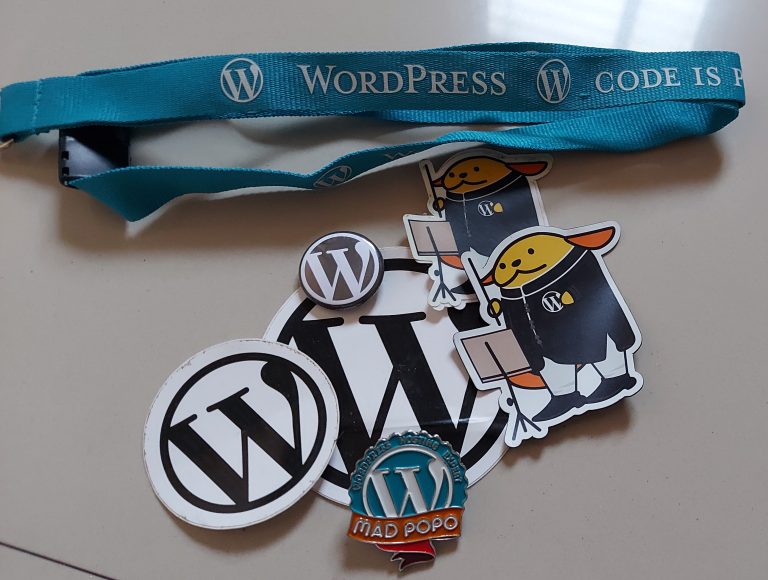 Several WordPress stickers, WordPress pins, Wapuu stickers as a music conductor, and a WordCamp lanyard.