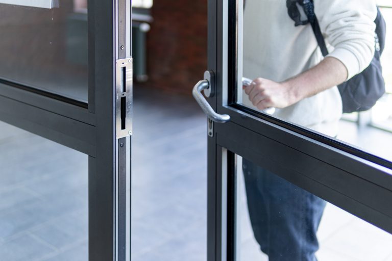 A man in a white shirt is using his left hand to open a set of aluminum-framed glass doors inside a building. His right hand is by his side. The man appears focused on the task, and his facial expression is neutral. The setting around the doors is not visible in the frame, and the lighting is relatively bright and uniform.