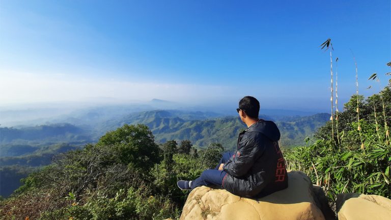 Person sitting on a rock on a mountain top looking over a vast expanse of forested mountains shrouded in clouds.? Sajek, Bangladesh.