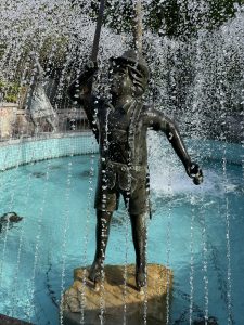 Statue of a small boy on old fashioned Lederhosen under the spray of a fountain.