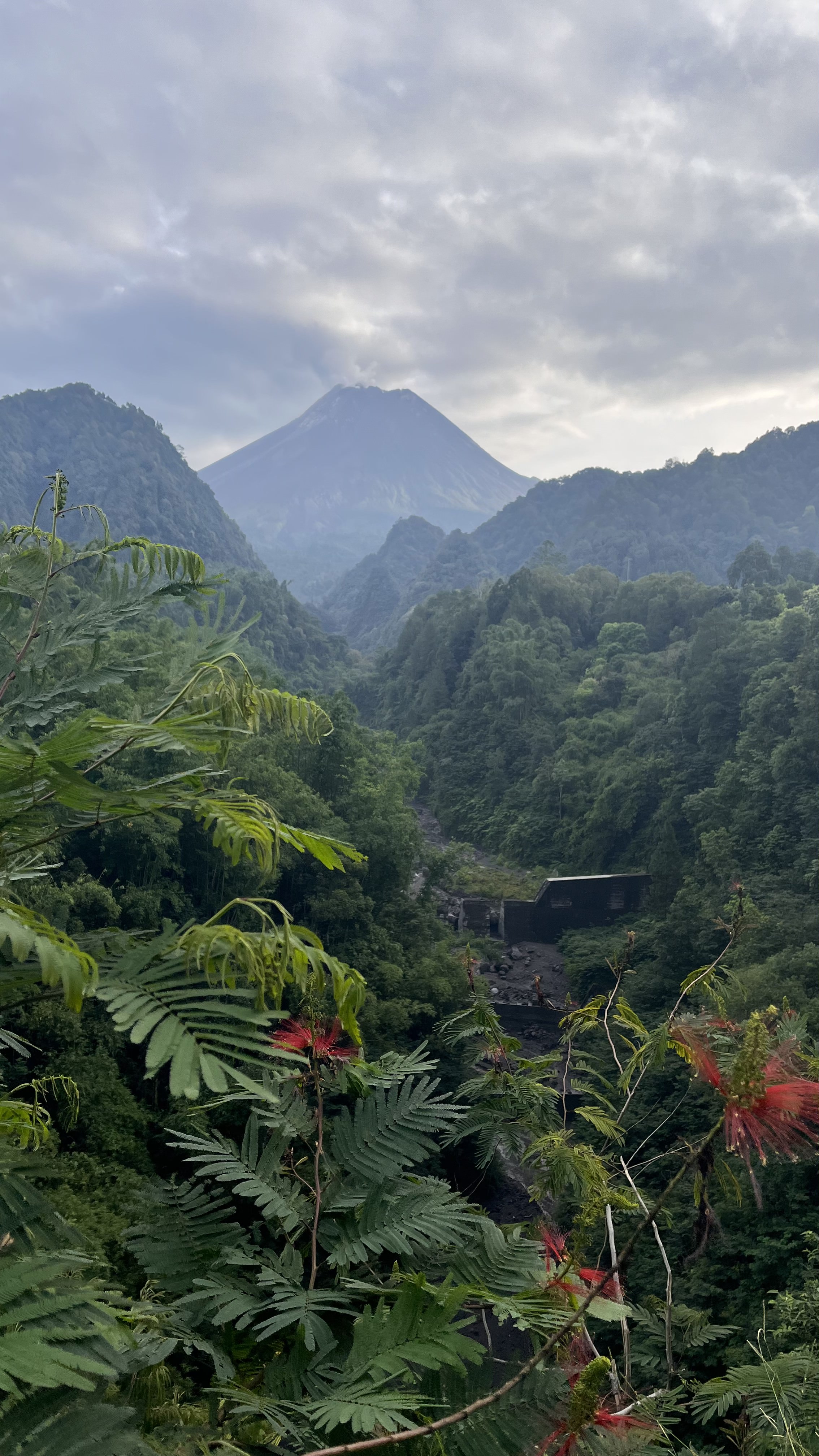 Photo of Mount Merapi volcano taken from Nawang Jagat camping area, Yogyakarta, Indonesia. View down a long valley filled with greenery, volcano on the horizon dominating the picture, smoke coming out the top.