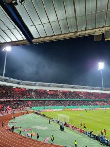 Interior of a football stadium at night. It is full of fans and the floodlights are turned on.