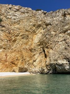 Golden Bay in Croatia, a towering rocky cliff with various shades of gold, brown and orange, overlooking a serene blue sea with a small pebbly beach visible at the base. 