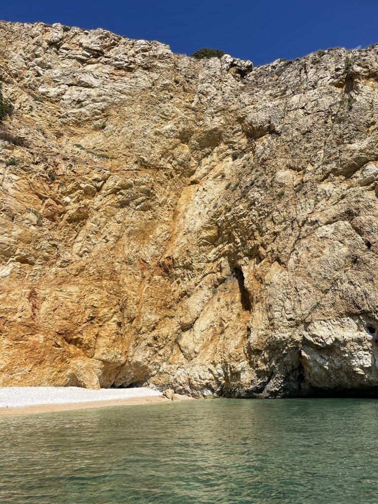 Golden Bay in Croatia, a towering rocky cliff with various shades of gold, brown and orange, overlooking a serene blue sea with a small pebbly beach visible at the base.