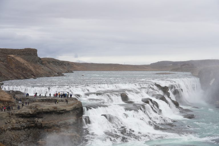 Very wide waterfalls, 100 feet or 30 meters tall, with a rock walkway on the left with 50 or 60 tourists looking on.