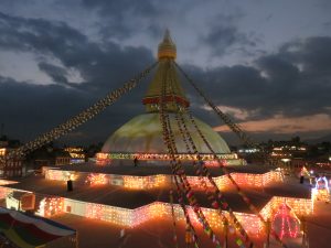 A night view of the Boudhanath Stupa. A temple with a peak in the center and prayer flags going from the peak to the corners of the building.  It's evening, and the building is covered in colored lights.