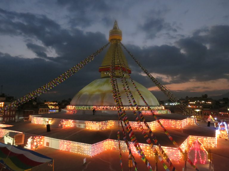 A night view of the Boudhanath Stupa. A temple with a peak in the center and prayer flags going from the peak to the corners of the building.? It’s evening, and the building is covered in colored lights.