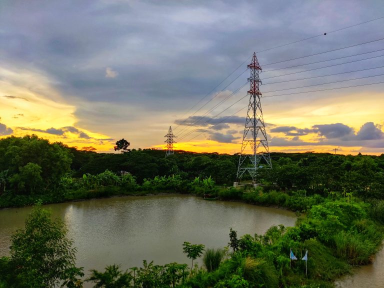 Looking down at a pond about 100 meters across.? Very large power lines from the right to the center horizon. Pond surrounded by thick forest. Center sky clouded, but horizon richly colored from the sunset.