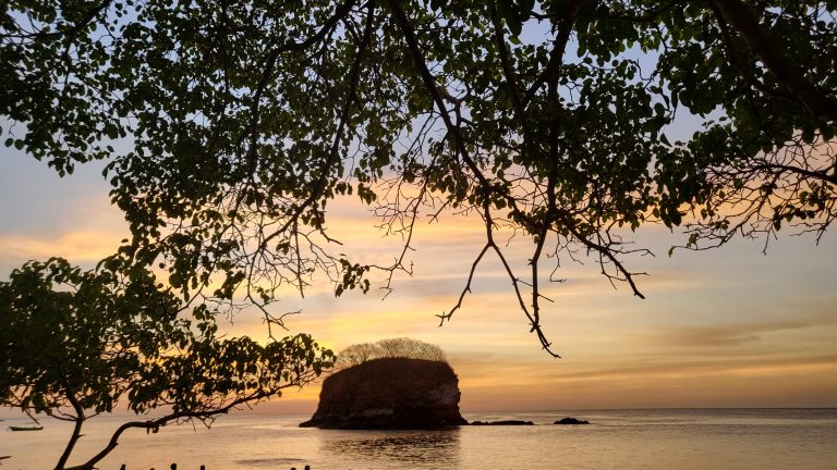 Playa, atardecer, mar. Looking through tree branches to an island just offshore.? The island has steep sides like a cake. The sun is setting behind the island.