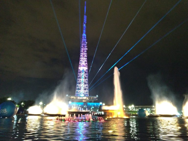 A tall TV tower in Brasília, Brazil, with purple lights and a fountain with brigh laser lights reaching the sky at night.