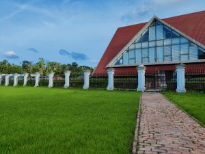 Liberation Museum Bhola, Bangladesh.? Grass lawn in the foreground, then a metal fence, and then an A frame building a couple stories high.