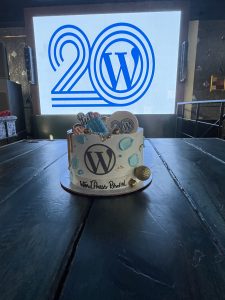 WordPress 20 anniversary cake. White cake on a table covered with WordPress buttons and swag with a large projection screen behind it with the WordPress 20th anniversary logo in it.
