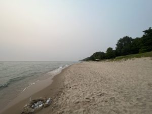 Lake Michigan shoreline on a foggy evening. Water on the left, trees and beach on the right. Small carn of rocks, sands, and gull feather in the foreground.