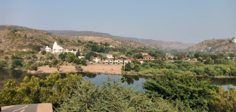 View across the countryside, a pond in the foreground, a small village on the other side. There’s foliage, but it looks a bit dry.