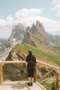 Person looking out at a viewpoint alongside a ridge of a rocky mountain with green grass on one side and cliffs on the other.