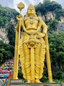 Lord Muruga aka Subrahmanya aka Kartikeya statue of Batu Caves temple. Kuala Lumpur, Malaysia.
Large statue made of something that looks like gold, mountains in the background, very large colourful staircase on the left with many people.