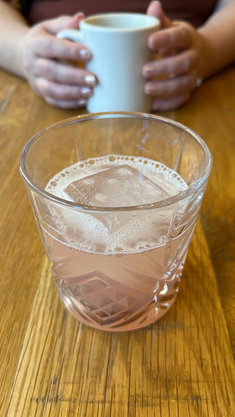 Agave Picante cocktail. Colors reminiscent of the 1920’s, large single ice cube. In the background, hands holding a coffee mug.