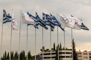 Greek and Olympic flags flying in the wind at sunset with apartment buildings and trees in the background
