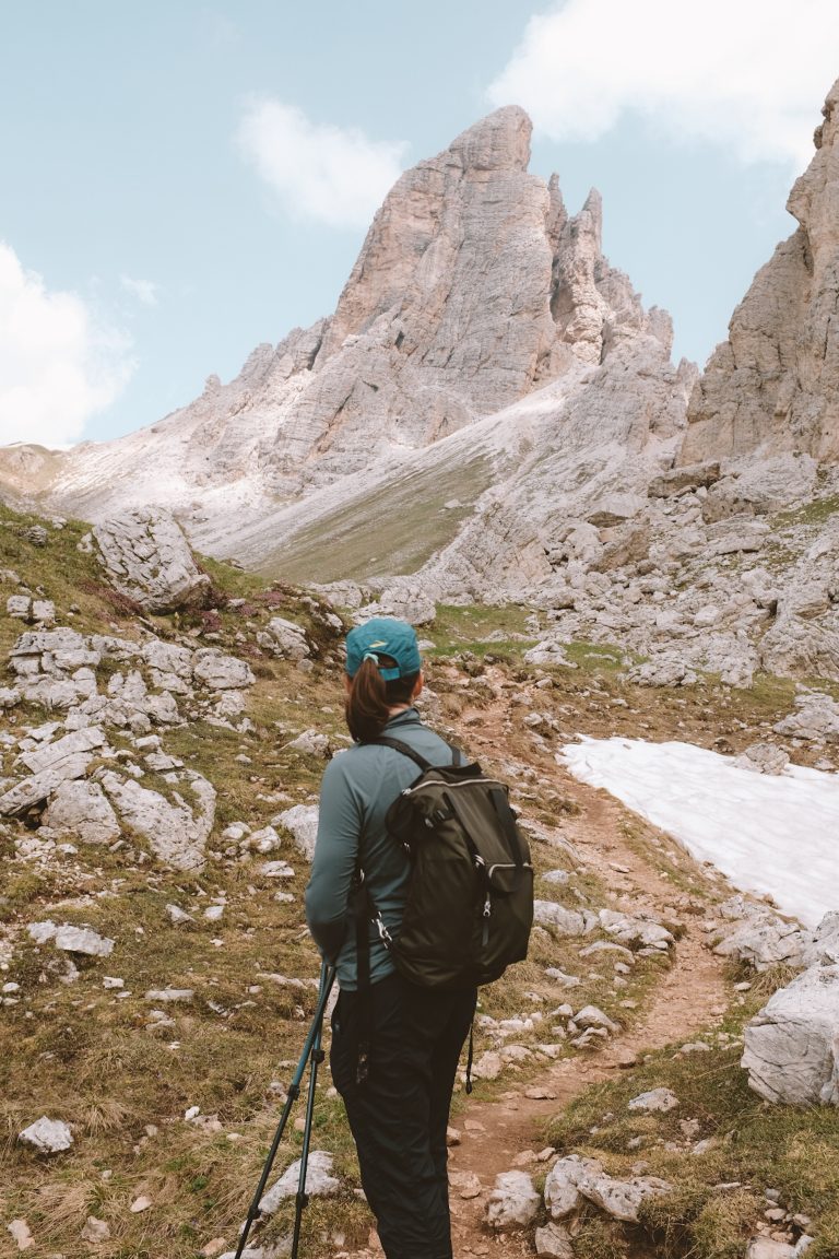 Person looking out at a trail in front of them with a rocky mountain peak above, rocky land to the left, and snow alongside the trail ahead to the right.