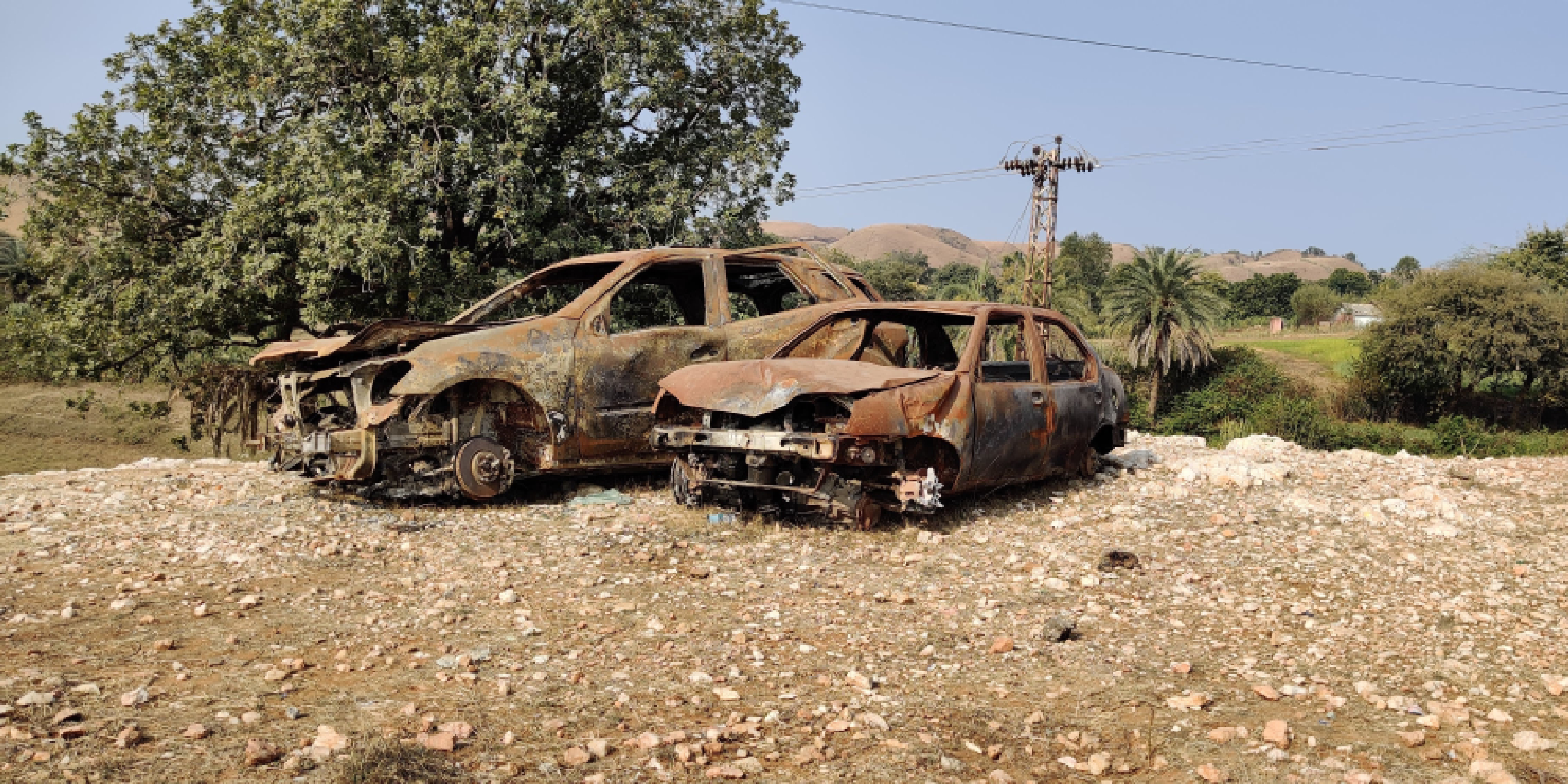 Two cars parked next to each other, both smashed and burned. Sitting on very rocky soil, a tree in the background.