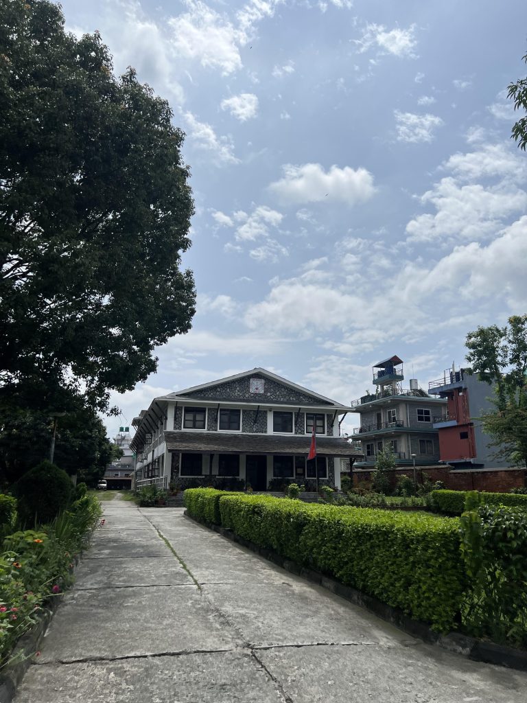 This picture shows a building built in Nepali architecture, surrounded by lush green trees in a compound. This is the headquarters of Annapurna Conservation Area Project, ACAP.