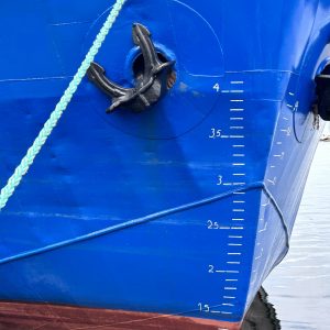 Close up of a blue ship’s plimsoll lines (reference marks located on a ship’s hull that indicates the maximum depth to which the vessel may be safely immersed when loaded with cargo). The anchor and a green rope is also visible.
