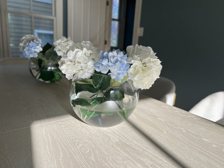 Hydrangeas in a globe vase sitting on a dining table with early morning sunlight stretching over them through a nearby window