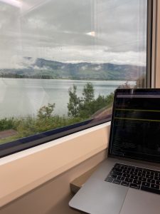 Travelling on the train with a laptop on a rainy day in Switzerland. A view out the window of a body of water with low clouds.  Laptop open on the table in the foreground.
