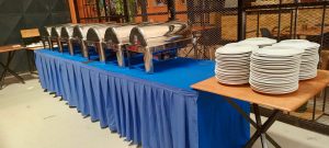 Hospitality mis-en-place, seven large stainless steel food storage containers sitting on a long table covered with blue material top and side as though ready for an event. A smaller table to the right has four piles of white plates stacked upon it. The set-up is indoor within a secure area formed by a padlocked diamond patterned metal fence through the fence we can an orange wall and smaller tables and chairs. Taken at Motiv, Kampala - Uganda