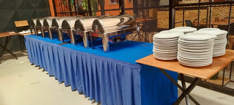 Hospitality mis-en-place, seven large stainless steel food storage containers sitting on a long table covered with blue material top and side as though ready for an event. A smaller table to the right has four piles of white plates stacked upon it. The set-up is indoor within a secure area formed by a padlocked diamond patterned metal fence through the fence we can an orange wall and smaller tables and chairs. Taken at Motiv, Kampala – Uganda