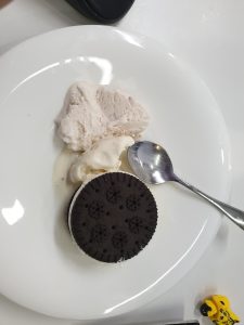 Vanilla ice cream on a plate with a spoon and a large chocolate cream sandwich cookie.  A small child’s toy to the side of the plate.
