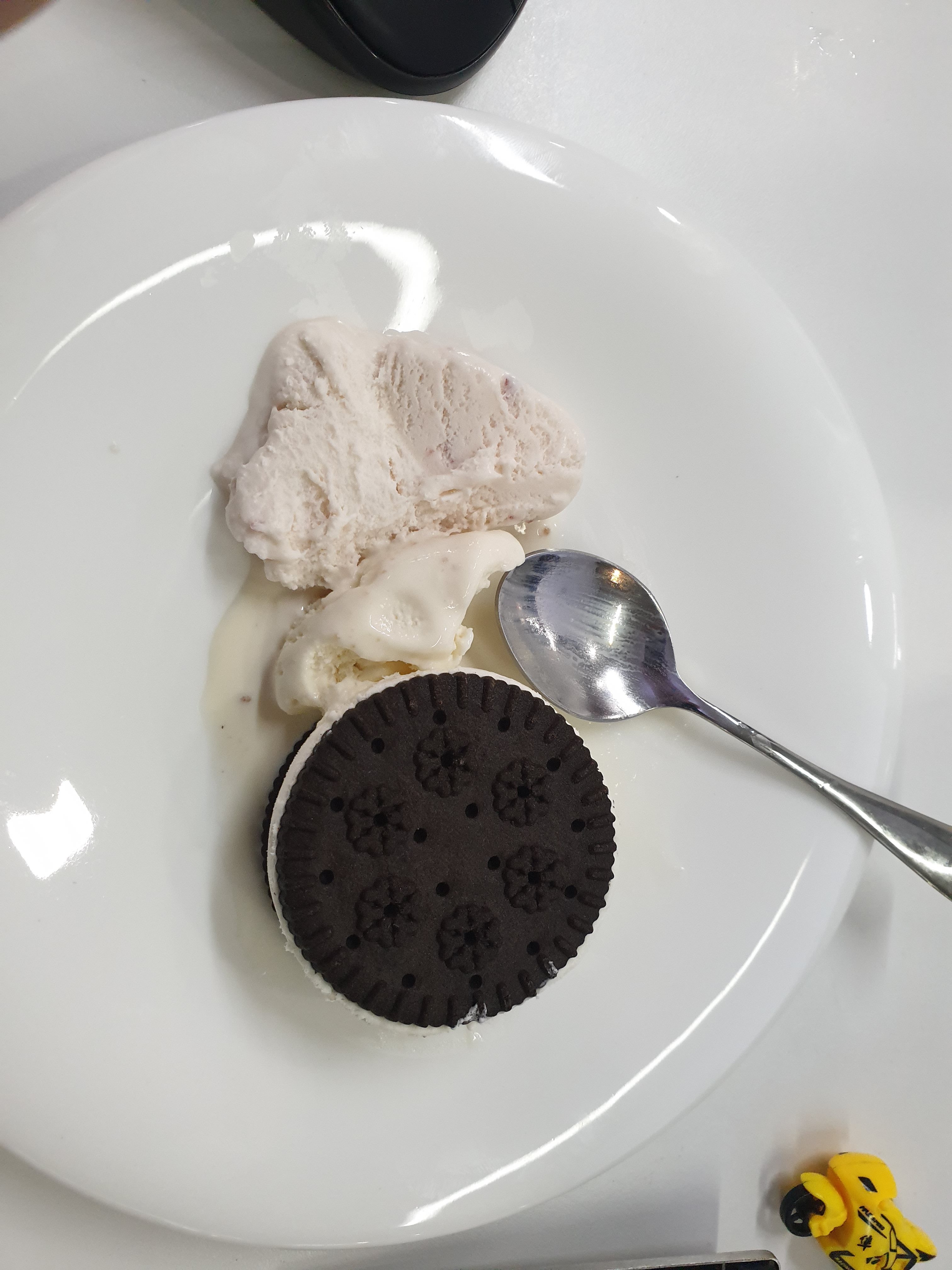 Vanilla ice cream on a plate with a spoon and a large chocolate cream sandwich cookie. A small child's toy to the side of the plate.
