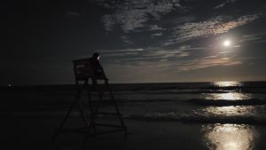 A lifeguard sits on his chair on a dark beach as the moon is setting. 
