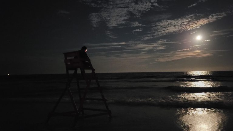 A lifeguard sits on his chair on a dark beach as the moon is setting.