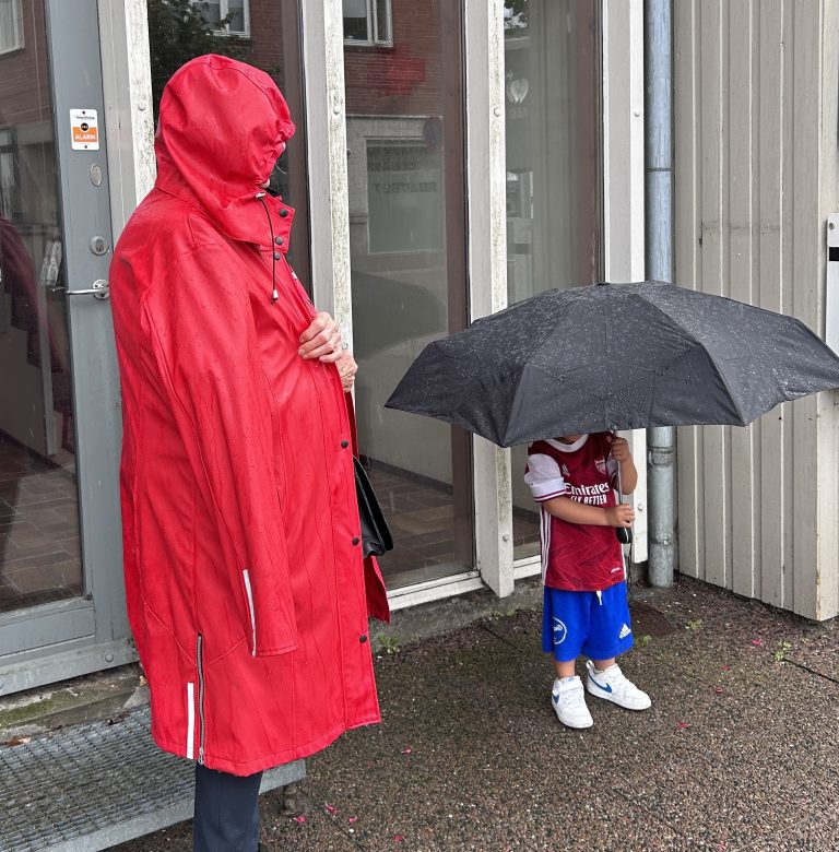 A child and an adult standing on a street in the rain. The adult is wearing a red raincoat and the child is wearing an oversized soccer outfit and holding a large black umbrella.