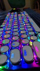 Closeup of a computer keyboard.  Each key is round, and has mirrored reflective sides. Each letter is backlit in a bright neon color.
