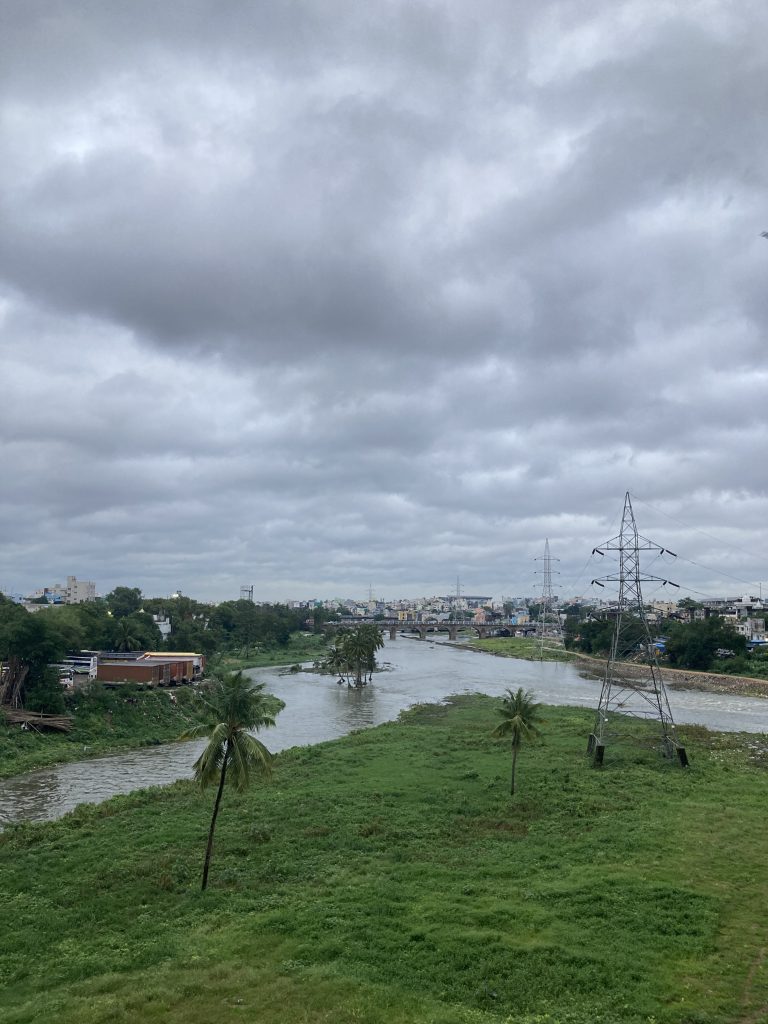 Two rivers merging at the edge of a city, going into the city.  Green grass and power lines in the foreground, city in the distance.