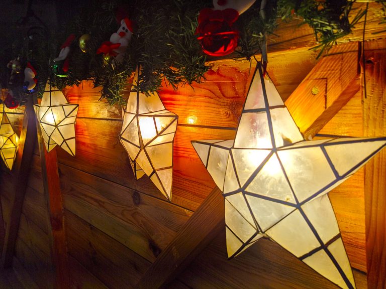 Four star shaped Christmas lanterns which have been constructed from triangular pieces of glass edged with metal. The lanterns are lit and hanging from a rustic wooden surface which has been topped with green tinsel and decorated with red, gold and green baubles, a small Santa decoration and a small snowman decoration.