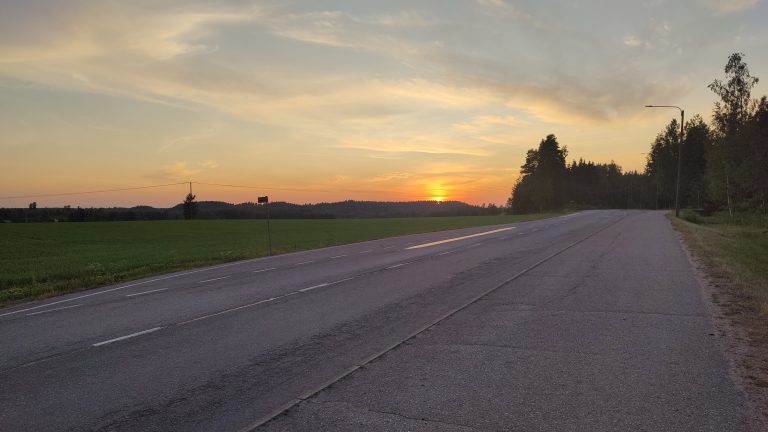 Asfphalt road, orange sunset, fields and forest in the distance.