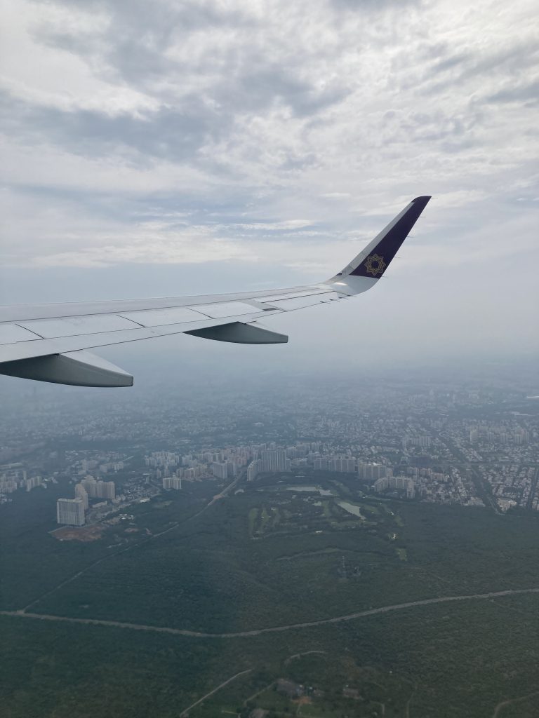 Looking down from a plane at a city on the ground. The wing of the plane is in view, with grey clouds.