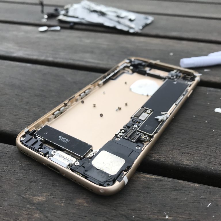 Part of an iPhone 7 which has been disassembled, the part we see is the inside view of the back of the phone. The beige casing containing some components including a battery and some tiny screws which would have held it together. it is lying on a rough wooden surface.