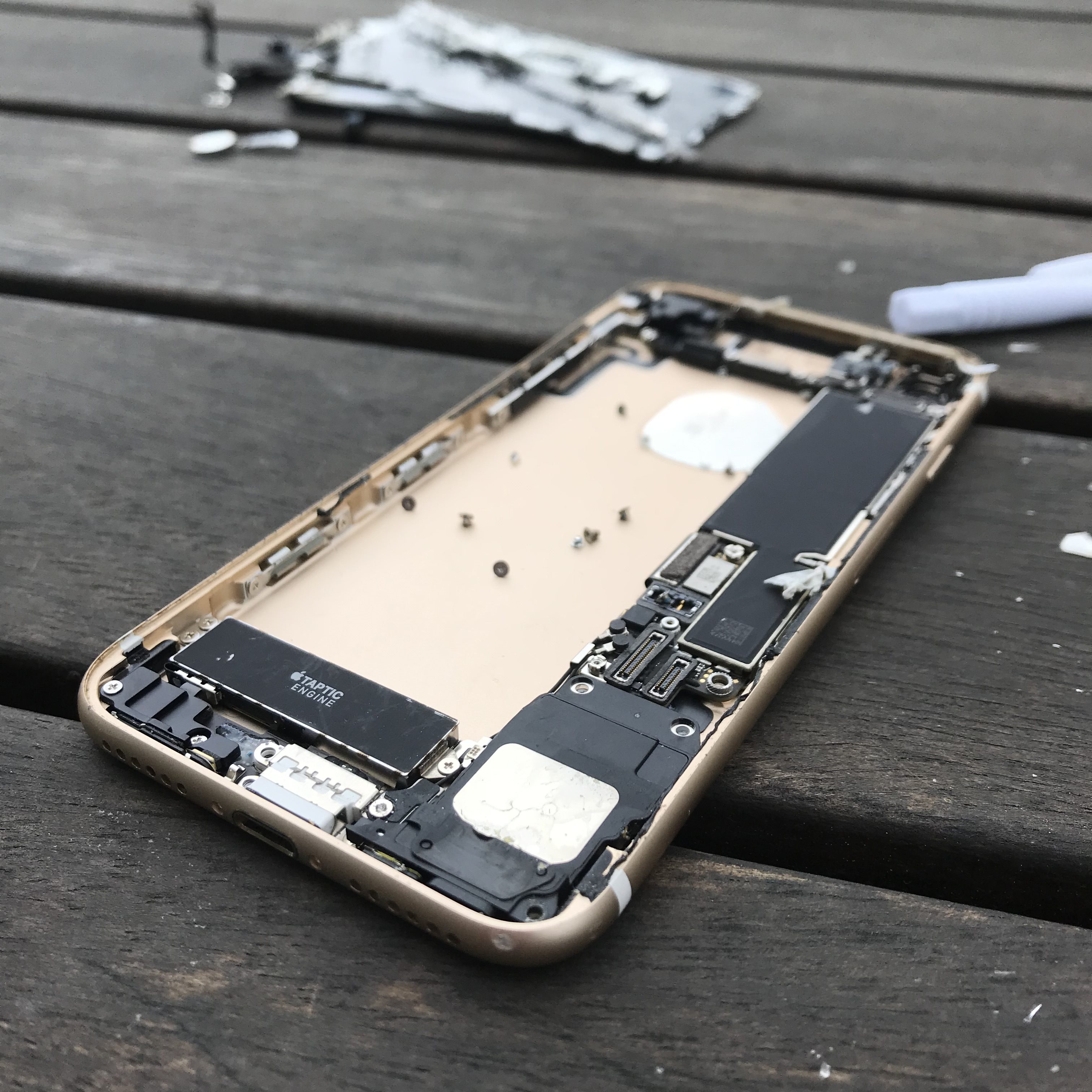 Part of an iPhone 7 which has been disassembled, the part we see is the inside view of the back of the phone. The beige casing containing some components including a battery and some tiny screws which would have held it together. it is lying on a rough wooden surface.
